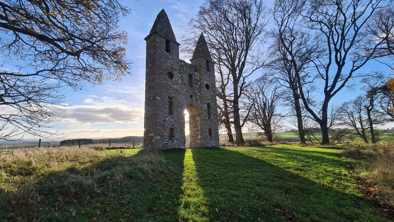 Historische stenen folly in tegenlicht met lange schaduwen over het gras, Hundy Mundy